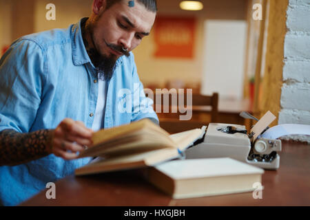 Portrait of stylish creative man with tattoos and beard turning pages of old book, while using old-school typewriter in quiet inspiring atmosphere Stock Photo