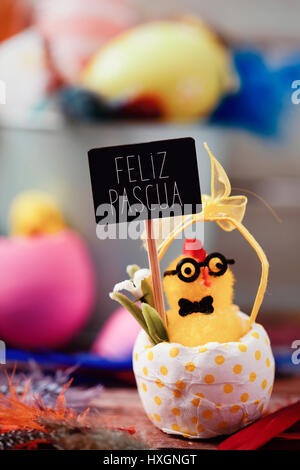 chick emerging from a cracked egg patterned with yellow dots holds a signboard with the text feliz pascua, happy easter in spanish, and a pile of deco Stock Photo