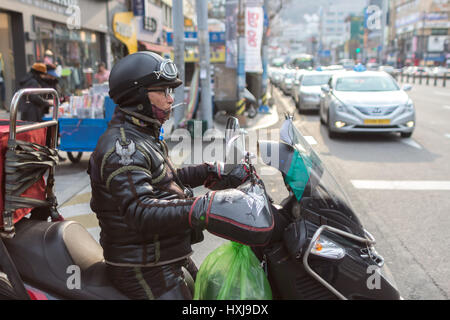 A man with a cigarette on his mouth driving a scooter motorcycle dressed with a Harley Davidson leather jacket in Busan, South Korea. Stock Photo
