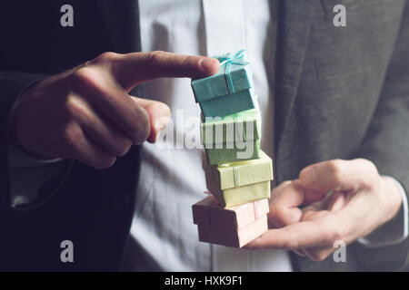 Horizontal close up of Caucasian man in black suit and white shirt holding with his index fingers three small colored gift boxes on top of each other  Stock Photo
