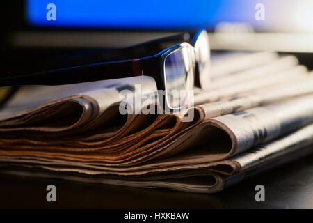 Glasses on stack of newspapers with blurred screen in background - News concept Stock Photo