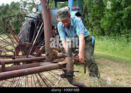 Lemozero, Olonets, Karelia, Russia - July 26, 2006: Farmer tractor driver, repairing ancient farm tractor rake in the fresh mown meadow. Stock Photo