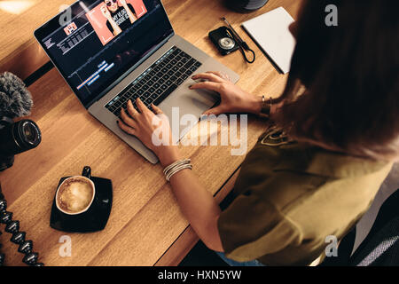 High angle view of female vlogger editing video on laptop. Young woman working on computer with coffee and cameras on table.