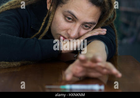 Man lying over table with dopey facial expression, reaching for syringe across desk, drug addiction concept Stock Photo