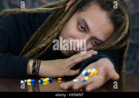 Man lying over table with dopey facial expression, colorful pills lying around desk, drug addiction concept Stock Photo