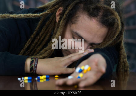 Man lying over table with dopey facial expression, colorful pills lying around desk, drug addiction concept Stock Photo