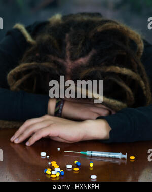 Man lying over table with dopey facial expression, colorful pills and syringe lying around desk, drug addiction concept Stock Photo