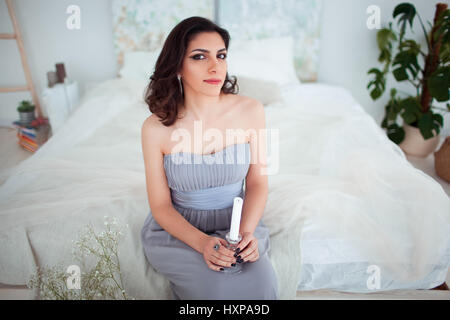 Cute girl with makeup in a blue evening dress sitting on the bed. In the hands holding a candle. Stock Photo