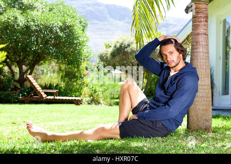 Full body portrait of young man with hand in hair sitting against tree Stock Photo