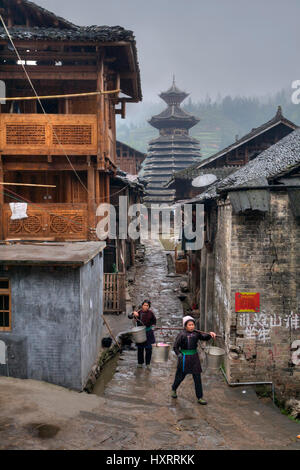 Zengchong Dong Village, Guizhou Province, China - April 13, 2010: Two women carry water on a yoke in rainy weather on the background drum tower of Don Stock Photo