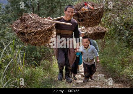 Langde Village, Guizhou, China - April 15, 2010: Chinese agricultural farm worker carries a yoke loaded, holding the hand of his son, a boy of about 5 Stock Photo