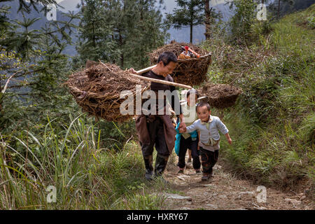 Langde Village, Guizhou, China - April 15, 2010: Peasants carrying a heavy load on their shoulders,  and holding the hand of a boy about 5 years old. Stock Photo