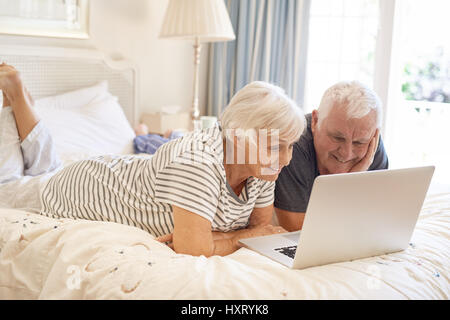 Happy senior couple lying on their bed using a laptop Stock Photo