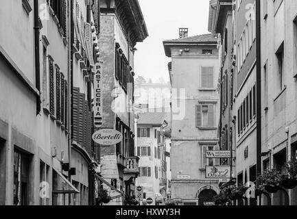 Trento, Italy - March 21, 2017: Houses in a narrow street in the old town with diverse facades, cafes and shop signs. The picture is monochrome. Stock Photo
