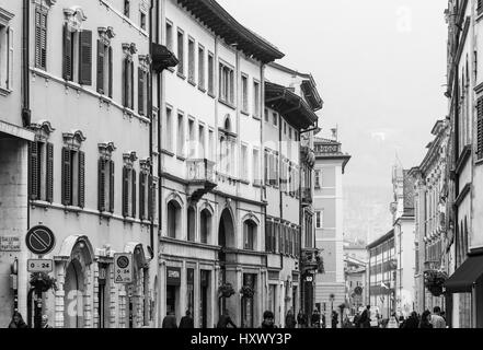 Trento, Italy - March 21, 2017: People in a shopping street in the old town with houses with diverse facades, cafes and shops. The picture is monochro Stock Photo