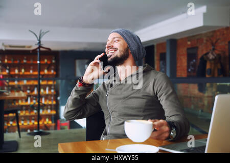 Young handsome hipster man with beard sitting in cafe talking mobile phone, holding cup of coffee and smiling. Laptop on wooden table. Stock Photo