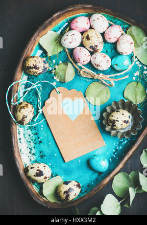 Colorful quail eggs, dried flowers and leaves for Easter holiday over turquoise blue tray with craft paper label in center with copy space. Easter gre Stock Photo