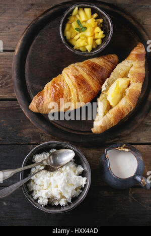 Breakfast with two croissant, butter, cottage cheese, cream and sliced mango fruit, served on wood round serving board over old wooden background. Top Stock Photo