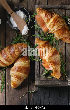Croissant with smoked salted salmon, spinach and arugula served with bowl of cream cheese over dark old wooden table. Top view Stock Photo