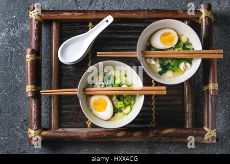 Two bowls with asian style soup with scrambled eggs, half of marinated egg, spring onion, spinach served with wood chopsticks and spoons on bamboo tra Stock Photo