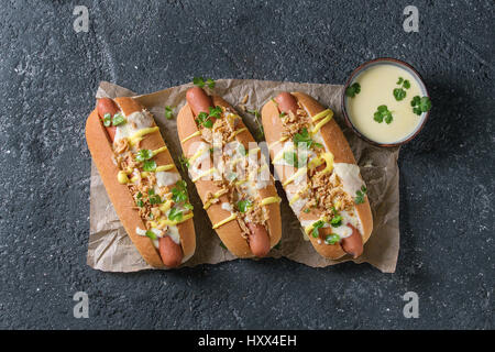 Hot dogs with sausage, fried onion, coriander leaves, cheese sauce and mustard, served on baking paper over black concrete texture background. Fast fo Stock Photo