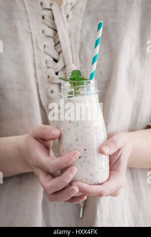 Bottle of smoothie with chia seeds, blueberries, mint leaves and retro striped cocktail tube in female hands. Woman in white linen dress. Healthy eati Stock Photo