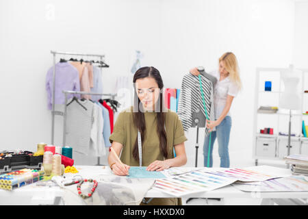 two young fashion designers collaborate for new clothing collection in their workshop , one in foreground sitting at the desk drawing and the other on Stock Photo