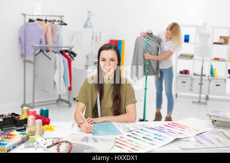 two young fashion designers collaborate for new clothing collection in their workshop , one in foreground sitting at the desk drawing and the other on Stock Photo