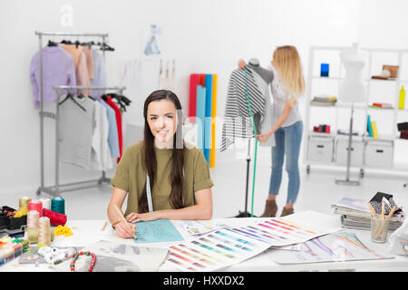 two young fashion designers collaborate for new clothing collection in their workshop , one in foreground sitting at the desk drawing and the other on Stock Photo