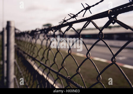 Chain-linked fence with barbed wire at top with grass and road in the background Stock Photo