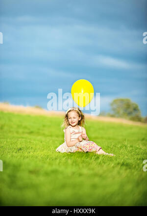 Little Blond Girl Sitting in the Countryside with a Single Yellow Balloon Stock Photo