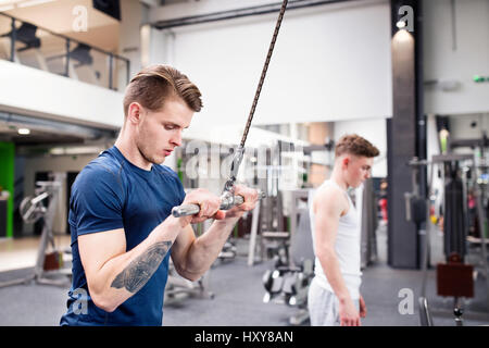 Young handsome fit man working out on pull-down machine in gym. Bodybuilder exercising with cable weight machine. Stock Photo