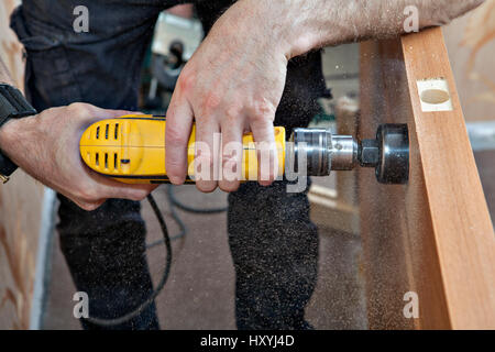 Hands woodworker with yellow electric drill, boring large hole of door lock, using hole saw drill bits, sawdust flying around, close-up. Stock Photo
