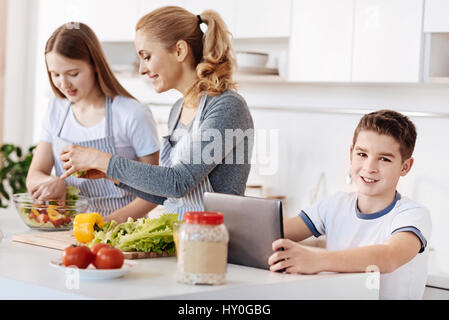 Modern technologies in use. Cheerful content smiling boy using tablet and standing in the kitchen while his mother and sister cooking in the backgroun Stock Photo