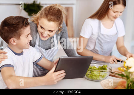 Share your emotions. Positive content mother talking with her son and using together tablet while cooking with the daughter in the kitchen Stock Photo
