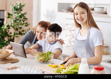 Involved in cooking. Cheerful content nice girl cutting vegetables and preparing salad while her mother and brother using tablet in the background Stock Photo
