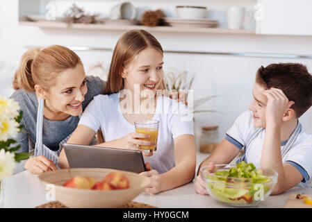 PLeasant time together. Pleasant smiling mother and her children using tablet and talking to each other while resting in the kitchen Stock Photo