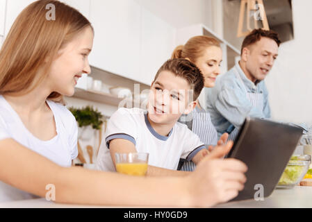 Lively talk. Positive smiling siblings talking and using tablet while their parents cooking for them Stock Photo