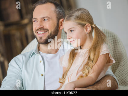 Smiling father and daughter sitting together in chair and looking away Stock Photo