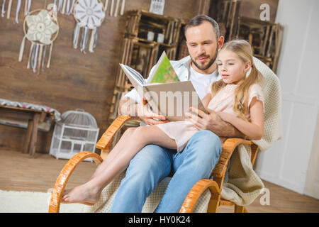 Father and daughter sitting together in rocking chair and reading book Stock Photo