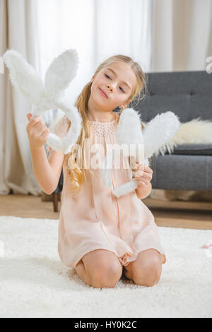 Pensive little girl sitting on white carpet and holding bunny ears Stock Photo