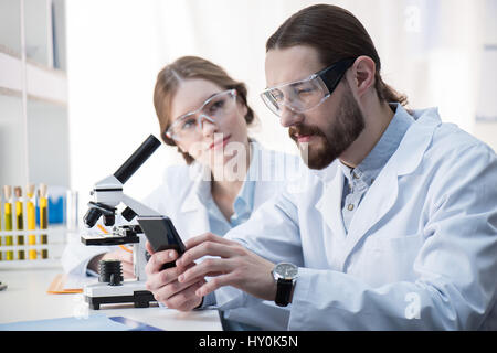 Young man chemist using smartphone  while working with female colleague in lab Stock Photo