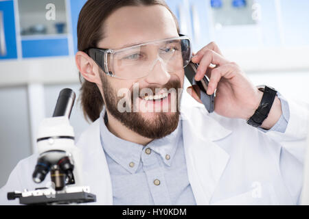 Happy young male scientist in protective glasses talking on smartphone Stock Photo