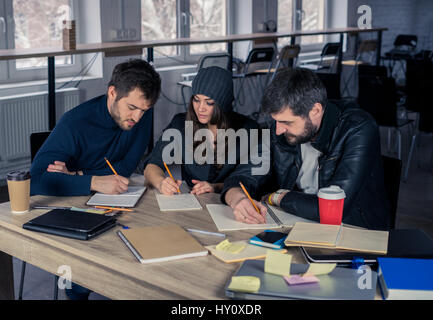 Young students writing in notepads or notebooks in lecture hall. Working environment with laptop, coffee, notepads and stationery. Stock Photo