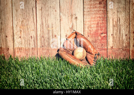 Old leather baseball mitt and ball on grass field against a rough wooden fence Stock Photo