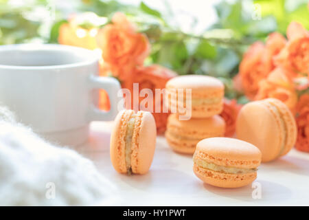 Orange mango or citrous macaroons, cup of coffee and fresh little roses on light wooden background. Sunlight. Coloring and processing photo with light Stock Photo