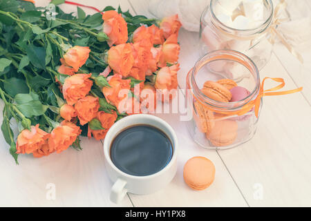 Orange mango macaroons, cup of coffee, marshmallow and fresh little roses on light wooden background. Sunlight. Coloring and processing photo with lig Stock Photo