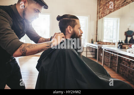 Hairdresser putting cutting hair cape on client at barbershop. Man with beard sitting at hair salon. Stock Photo