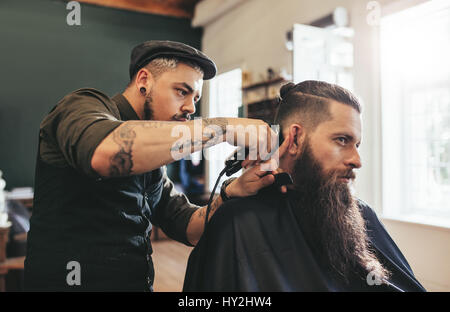 Handsome young bearded caucasian man getting trendy haircut in barbershop. Hairstylist serving client in salon. Stock Photo