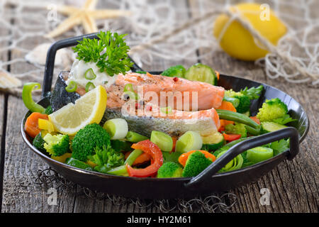 Tasty fried and grilled salmon steak on mixed colorful vegetables served in a frying pan,  lemons and a fishing net in the background Stock Photo